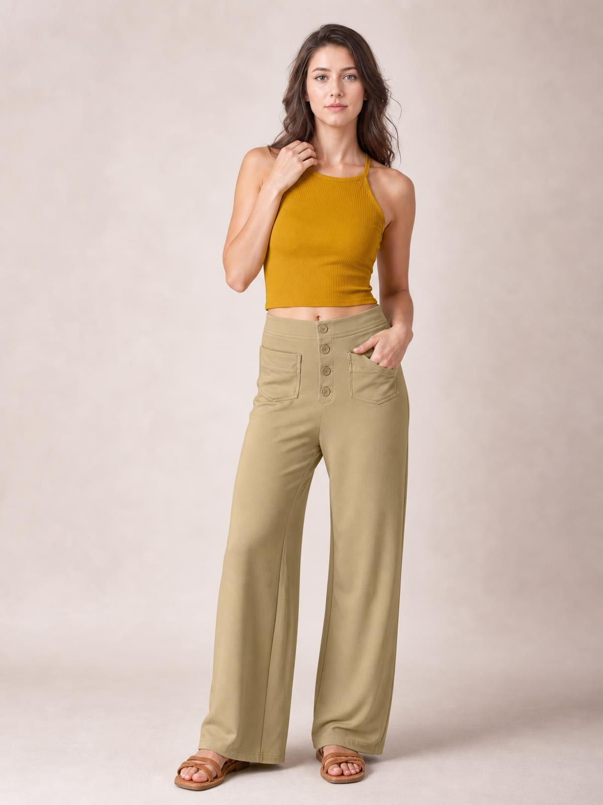 A standing model wears tan high-waist wide-leg pants with a front button closure and patch pockets, placing one hand in a pocket to show the relaxed silhouette.