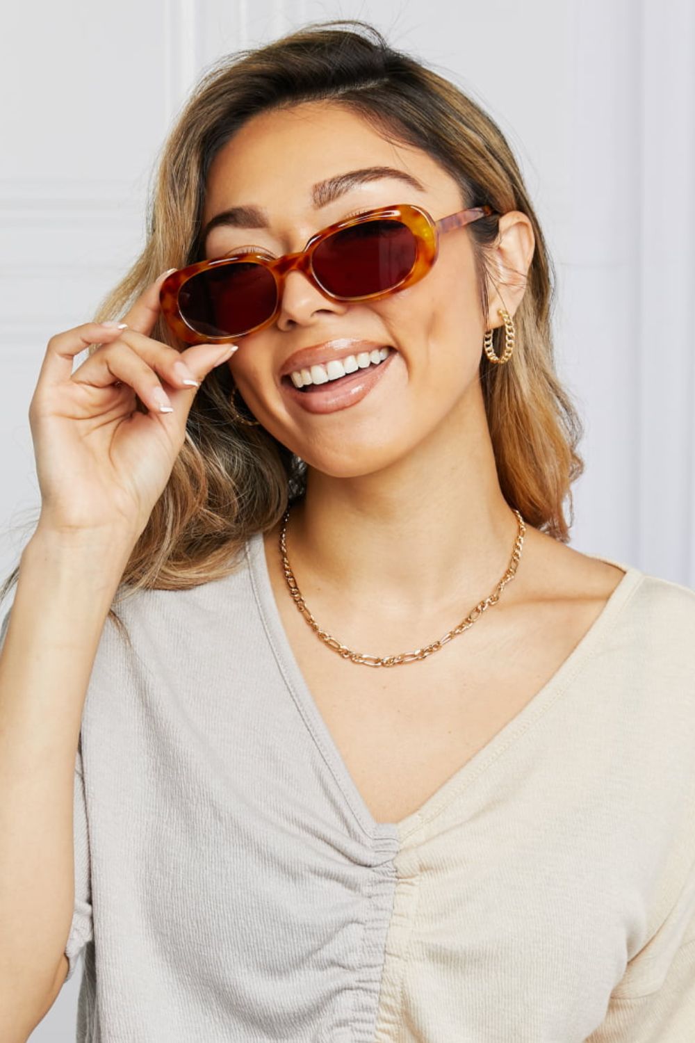 A smiling model adjusts amber tortoiseshell oval full-rim sunglasses with dark lenses, showcasing their softly curved vintage-inspired silhouette.