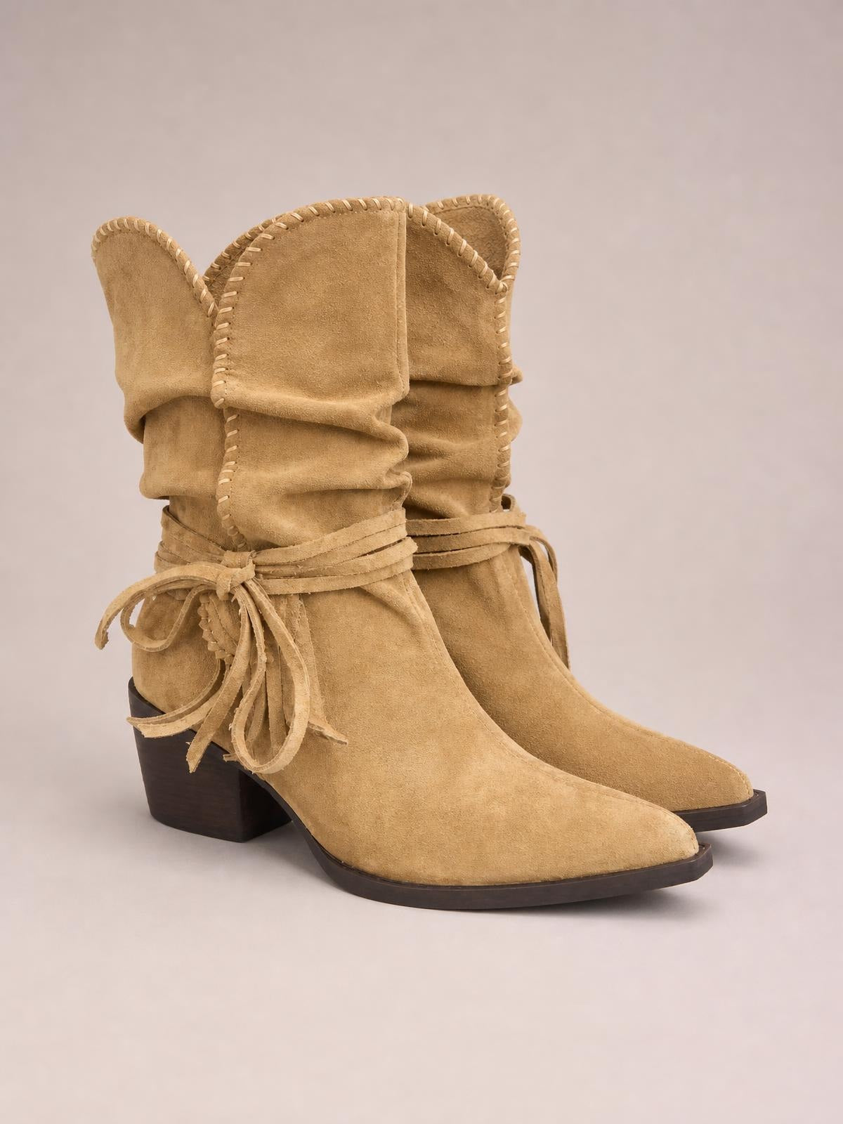 Tan suede point-toe block-heel boots feature a slouchy shaft, wraparound tie details, decorative stitching, and sturdy rubber soles.