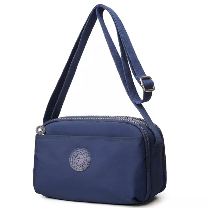Small navy nylon crossbody bag with an adjustable strap, zip-top closure, front slip pocket, rounded silhouette, and compact structured construction.