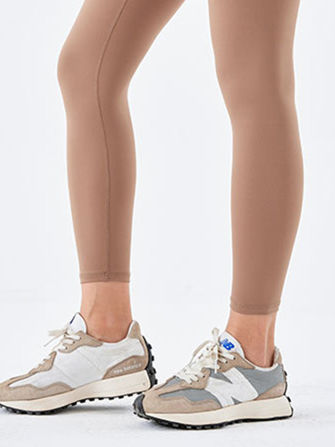 A model wears tan, ankle-length sports leggings with a close, stretchy silhouette and visible stitching along the legs while standing in sneakers.