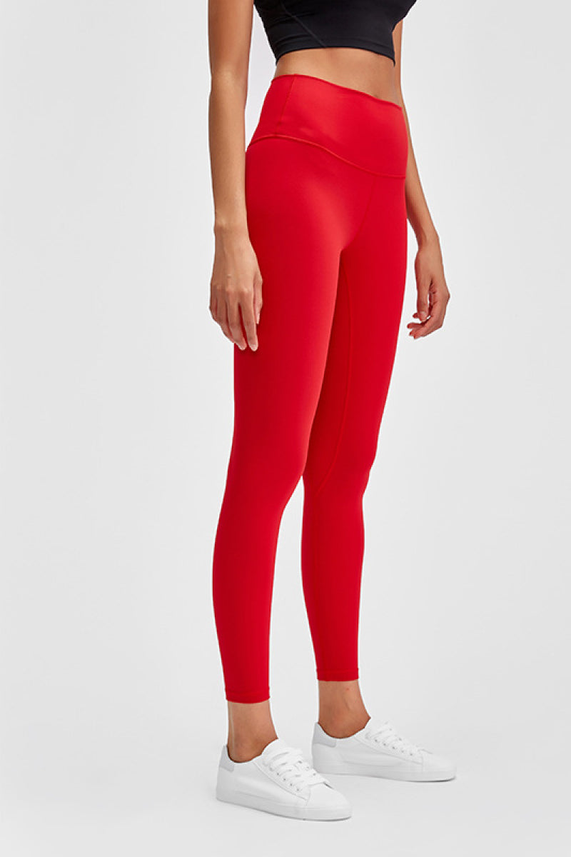 Model stands in bright red sports leggings with a wide seamless band waist, fitted ankle-length silhouette, and smooth basic construction.