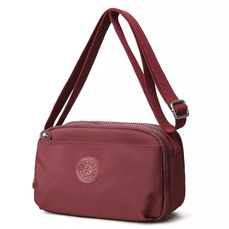 Small burgundy nylon crossbody bag with an adjustable strap, compact rectangular silhouette, rounded corners, front slip pocket, and top zipper closure.