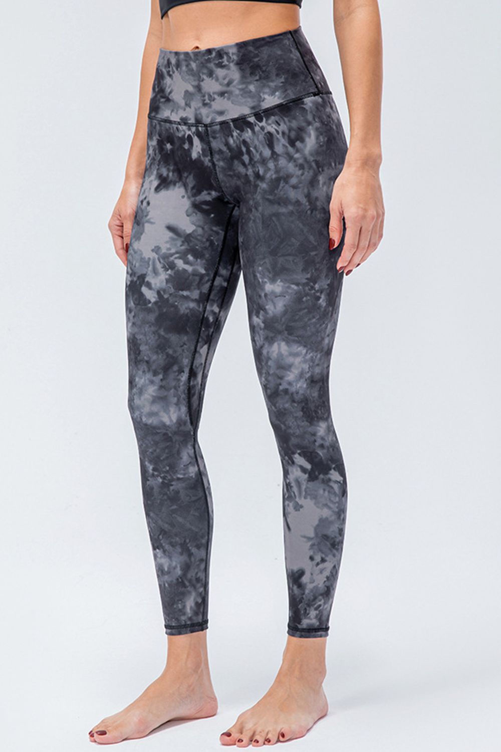 Model standing in dark gray tie-dye active leggings with a wide waistband, slim ankle-length silhouette, and visible front seam construction.