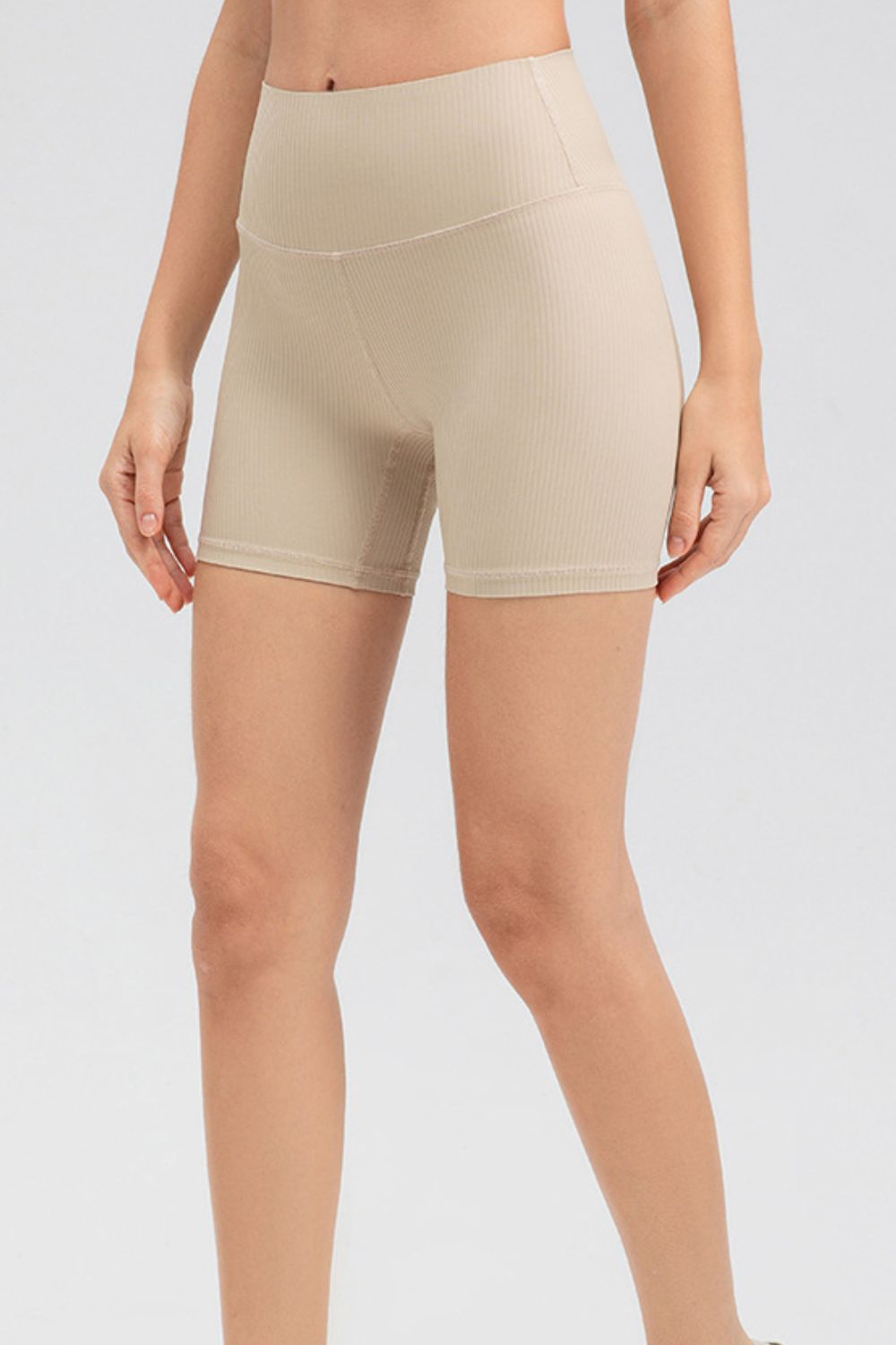 A model wears solid beige slim-fit sports shorts with a wide waistband, smooth stretch construction, fitted mid-thigh silhouette, and clean seamed hems.