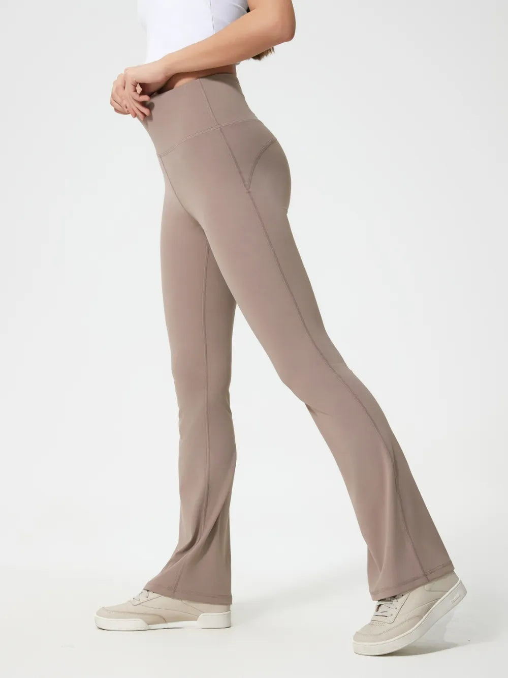 A model poses in taupe high waist active pants with a close-fitting, flared-leg silhouette, wide waistband, and visible contour seams.