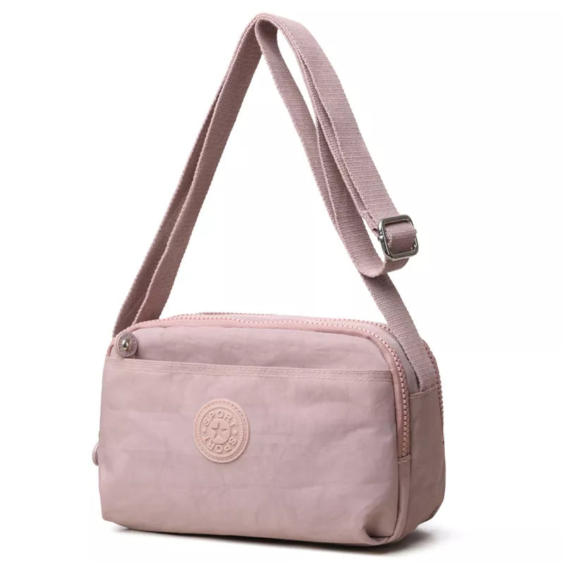 Small pink nylon crossbody bag with an adjustable strap, rectangular silhouette, zippered top, and front circular logo patch.