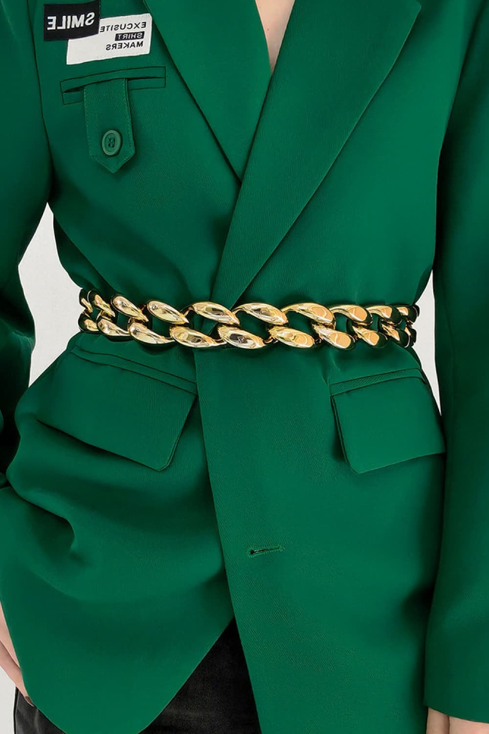 A model wears a gold-tone acrylic curb chain belt with bold interlocking links around the waist of a tailored green blazer.