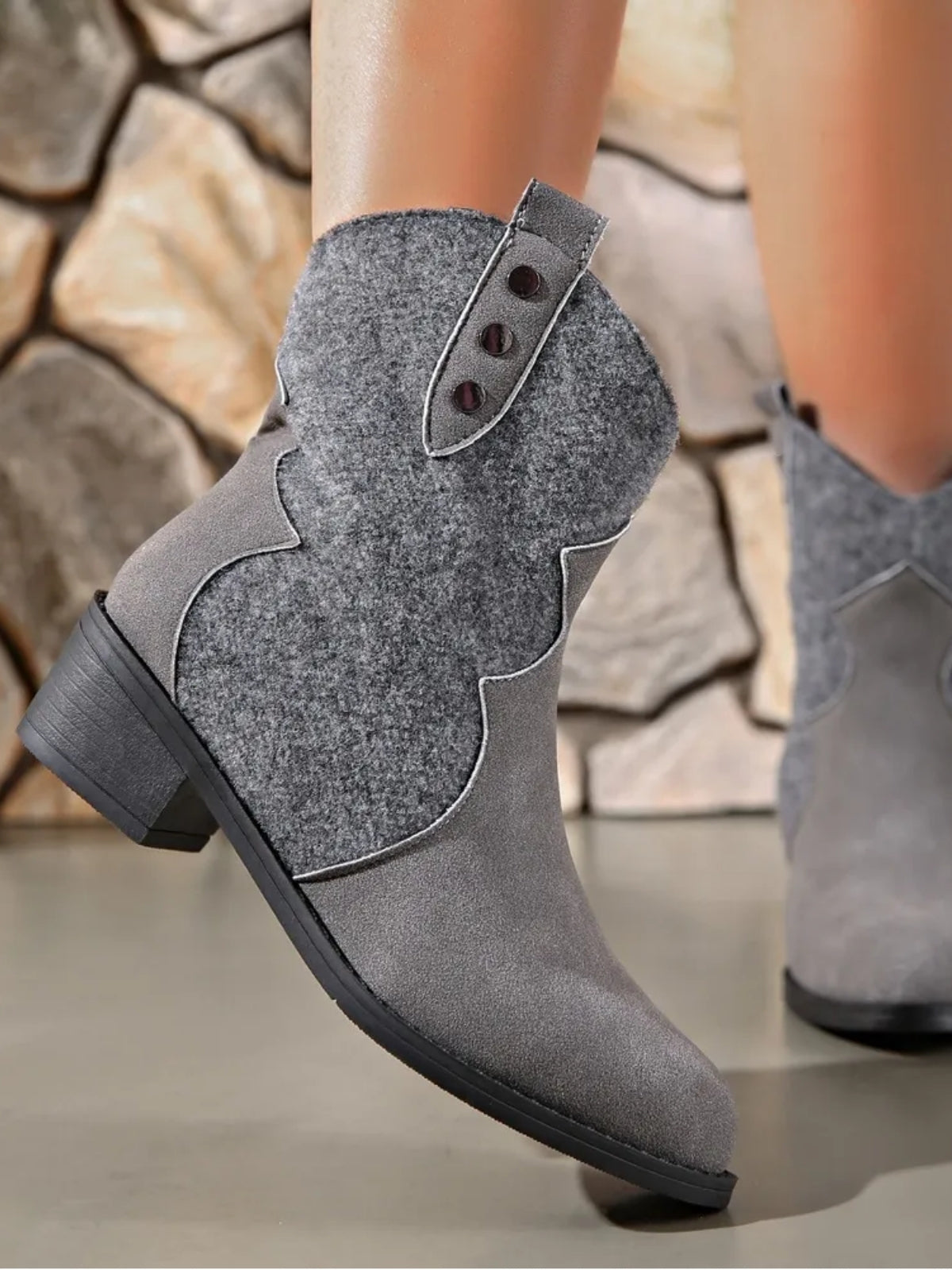 A model lifts one foot to show a gray suede ankle boot with a pointed toe, low block heel, contrast piping, and a decorative buttoned upper panel.