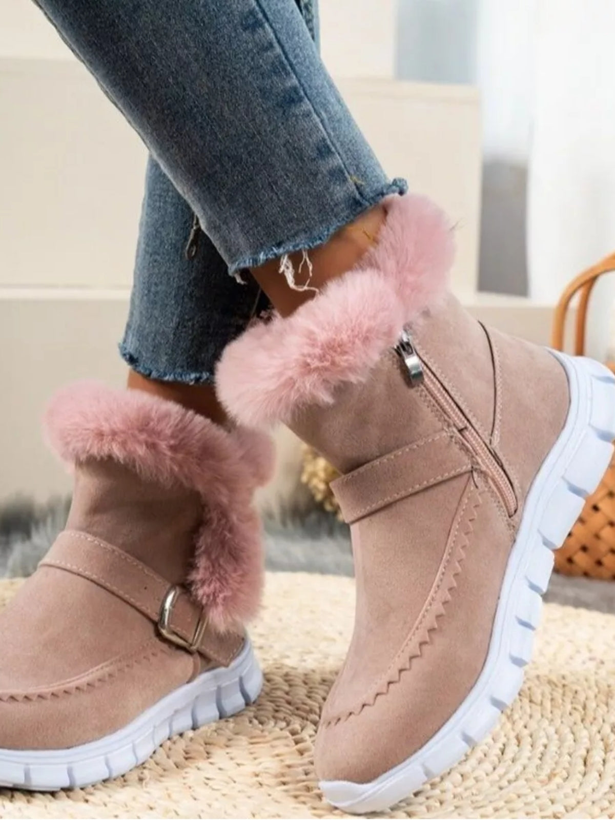 A model poses with crossed legs in taupe suede buckle boots featuring soft pink fuzzy trim and chunky white rubber soles with a flat silhouette.