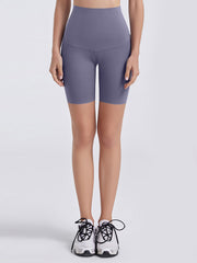 Model wears purple wide-waistband sports shorts with a high-rise, fitted mid-thigh silhouette and curved front seam, standing with hands at her sides.