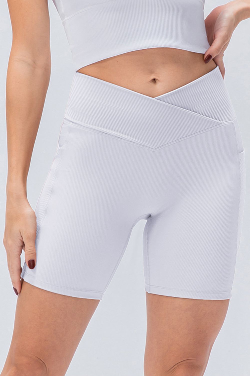 A standing model wears solid white slim-fit sports shorts with a crossed V-waistband, stretchy fitted silhouette, and clean stitched hems.