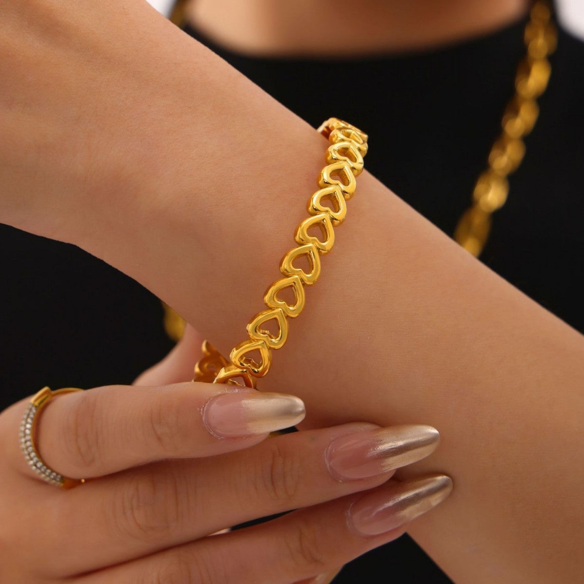 A close-up shows a hand touching a wrist wearing an 18K gold-plated stainless steel bracelet with linked heart-shaped charms.