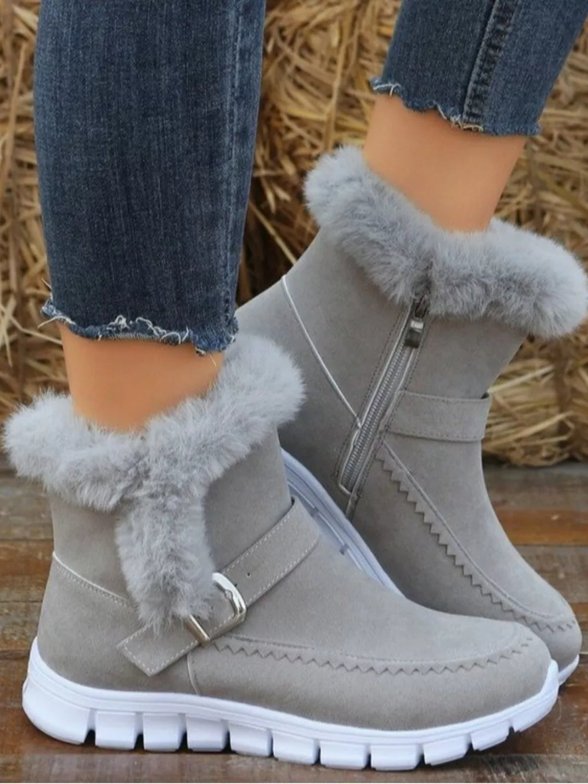 A model wears light gray suede flat boots with fuzzy trim, buckle straps, side zippers, decorative stitching, and chunky white rubber soles.