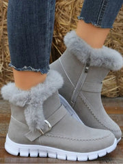 A model wears light gray suede flat boots with fuzzy trim, buckle straps, side zippers, decorative stitching, and chunky white rubber soles.
