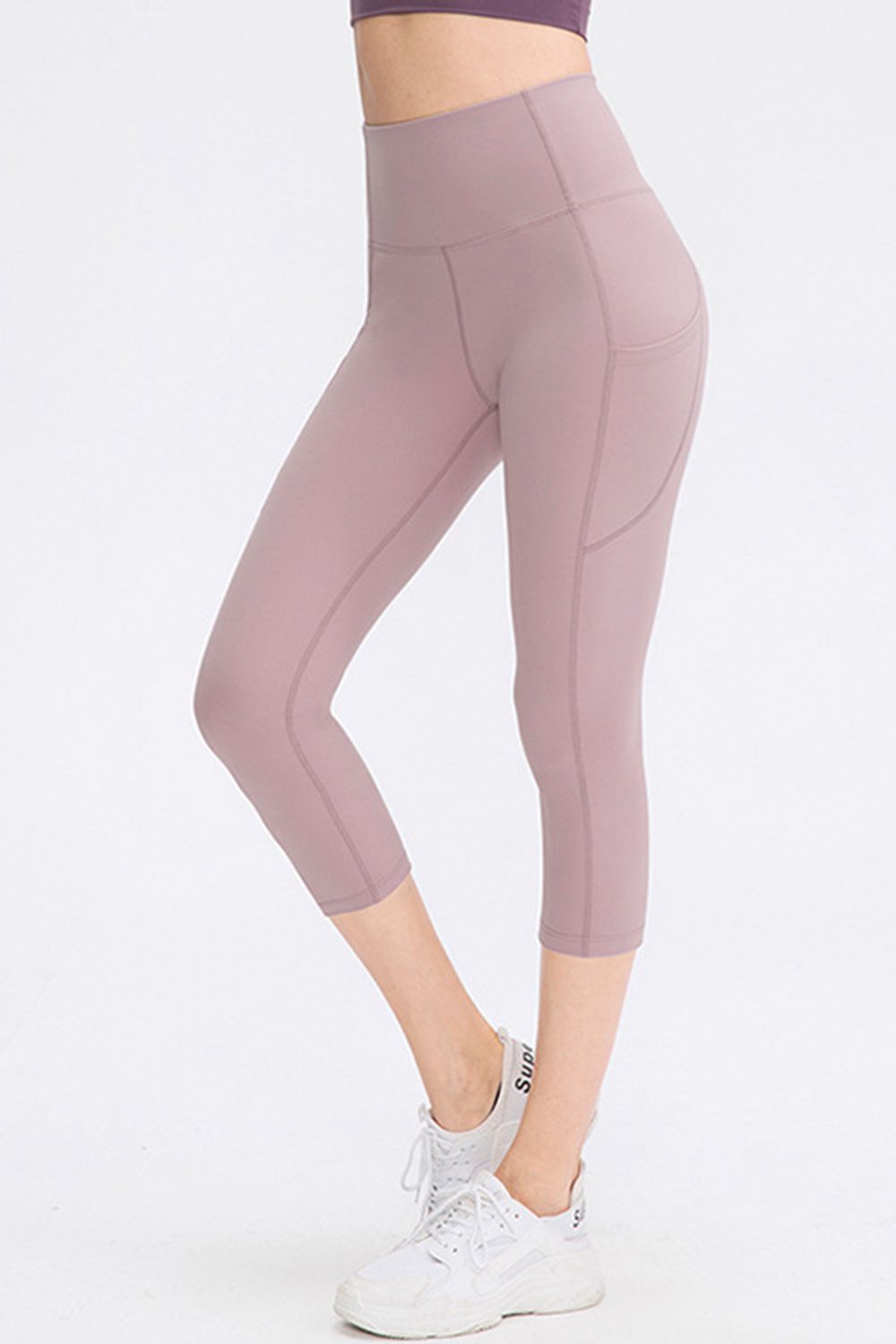 A model poses in dusty mauve cropped active leggings with a wide waistband, slim silhouette, visible pocket, curved seam construction, and one bent leg.