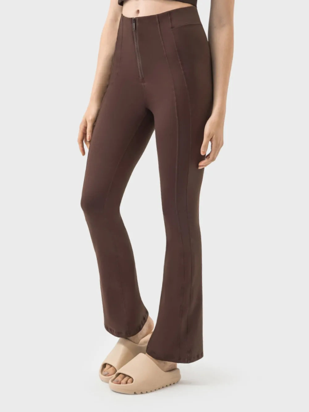 A model wears brown high-waist active pants with a front zipper, vertical seam detailing, a slim fit through the thighs, and subtly flared hems.