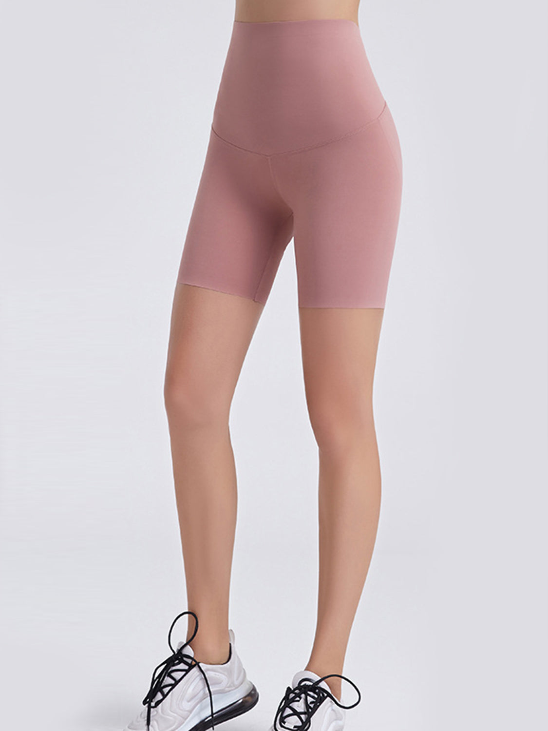 A model wears pink wide-waistband sports shorts with a smooth, fitted mid-thigh silhouette and curved front seams, standing with feet apart.