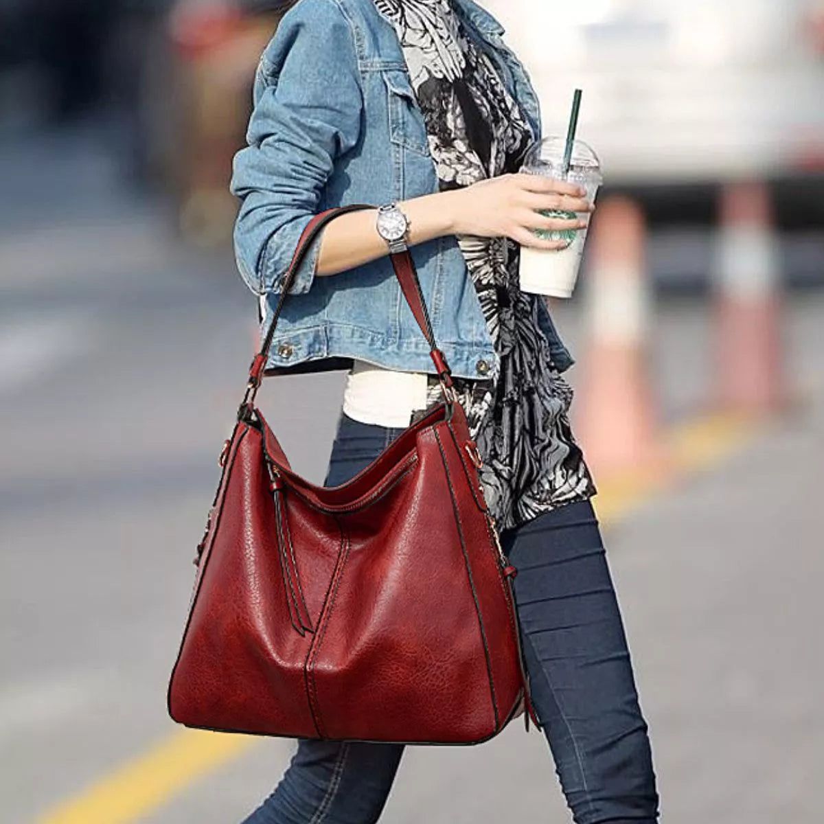 A model walks with a medium red PU leather shoulder bag, showing its slouchy silhouette, twin straps, top opening, and visible stitched construction.