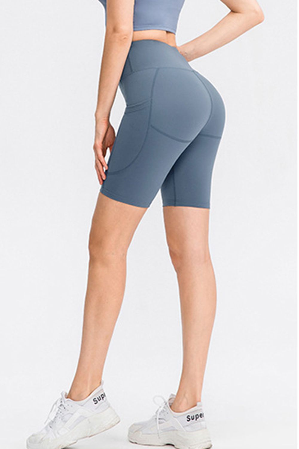 Model in a rear three-quarter pose wears solid slate-blue sports shorts with a wide waistband, fitted biker-length silhouette, side pocket, and seam detailing.
