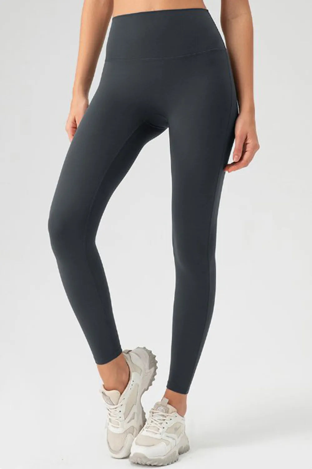 Model wears dark gray high-waist skinny active pants with a smooth, stretchy fitted silhouette and ankle-length construction, standing with one leg crossed forward.