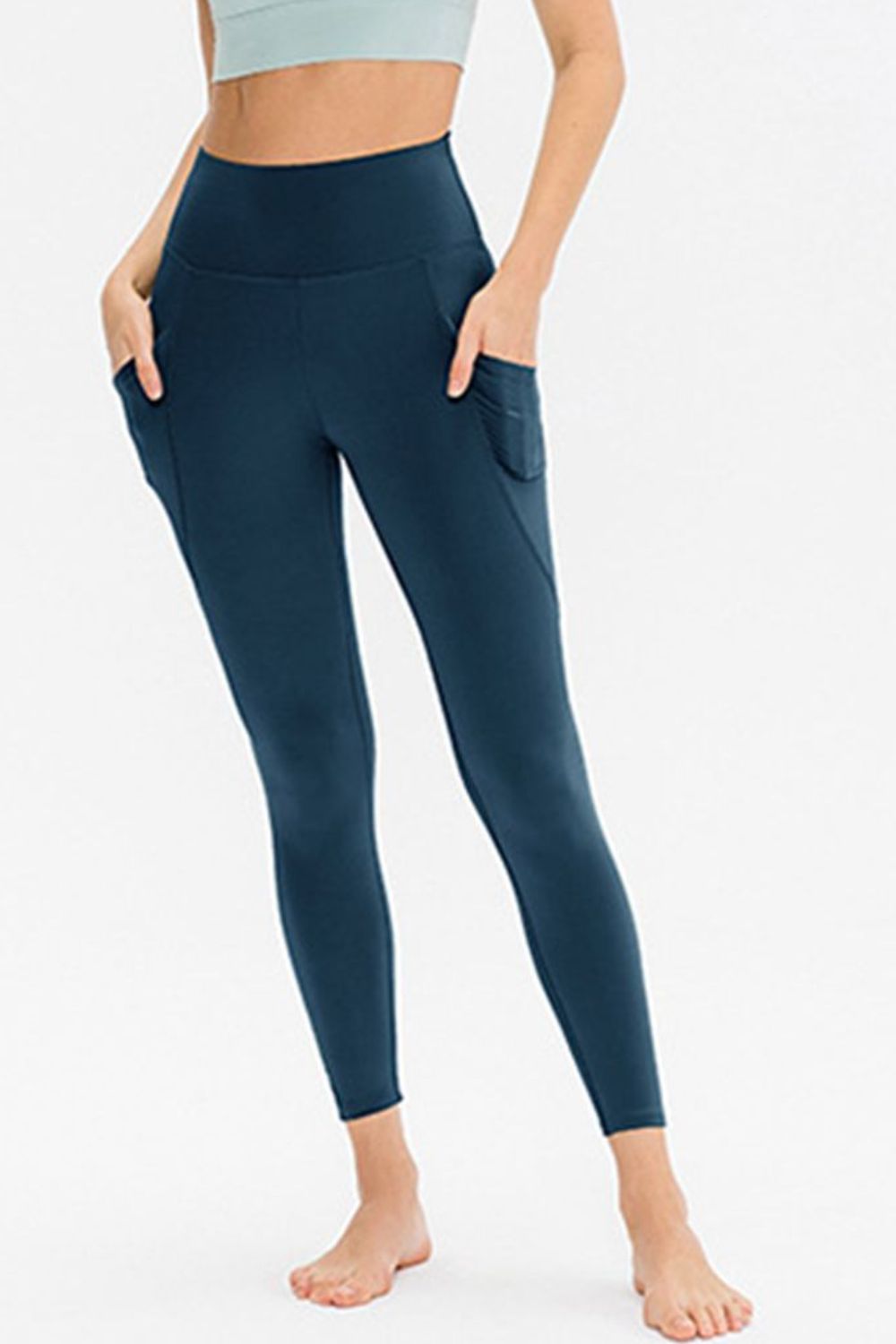 Model wears solid navy slim-fit active leggings with a high waistband, long tapered legs, and side pockets, standing with hands resting near the hips.