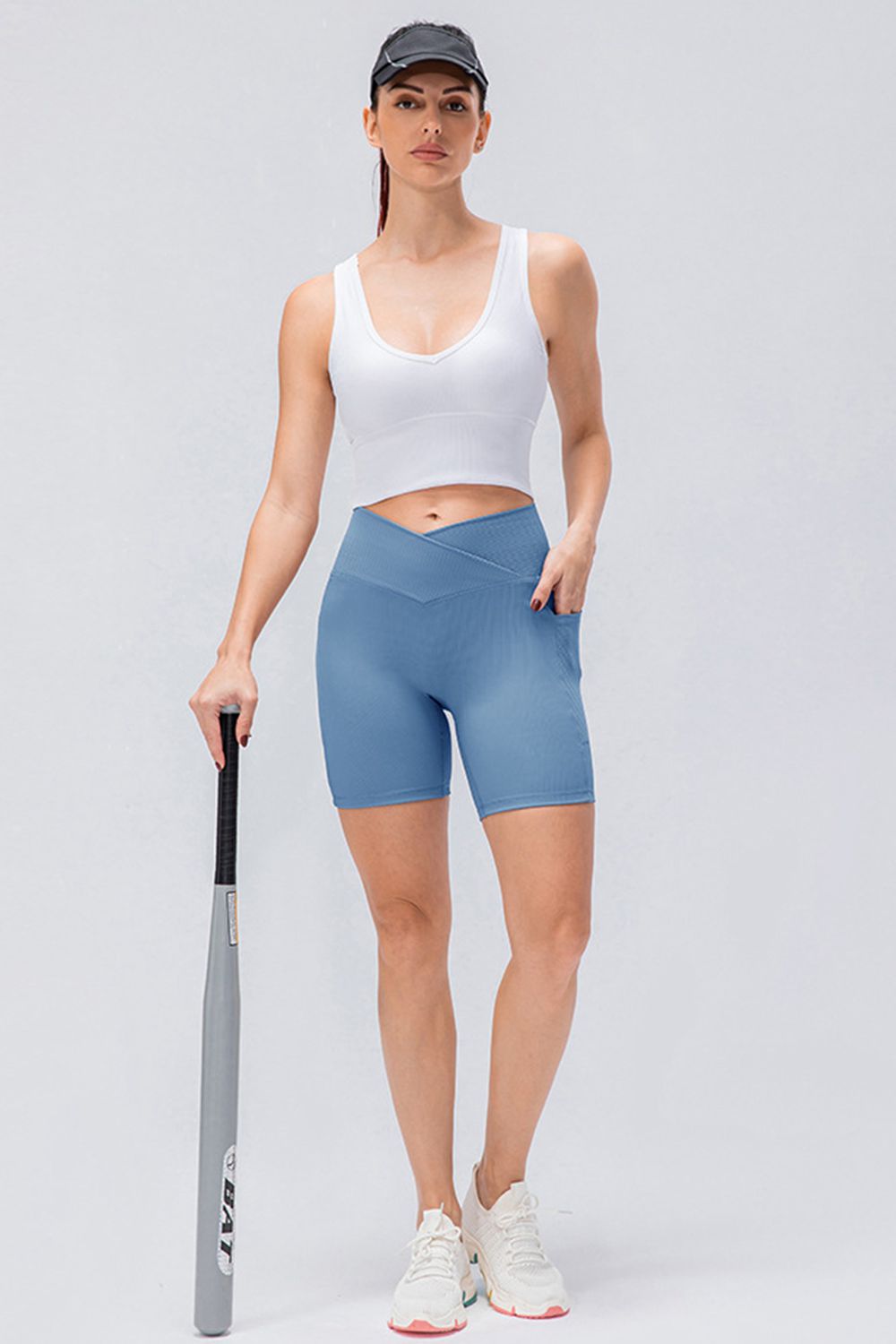 Model wearing blue slim-fit sports shorts with a V-waistband and mid-thigh silhouette, standing in an athletic pose while holding a bat.