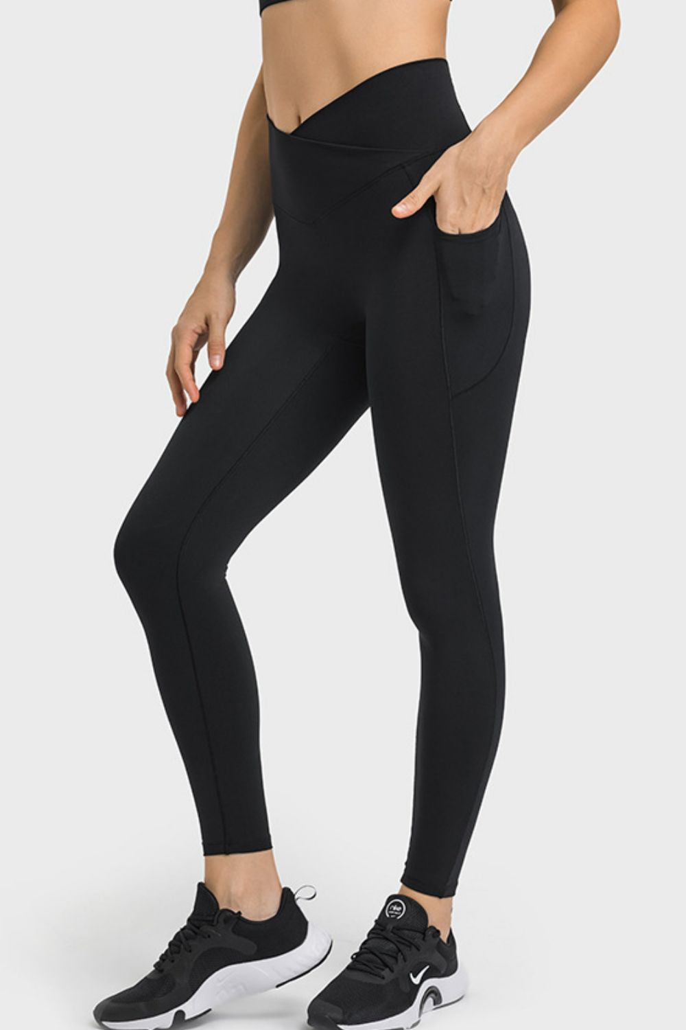 A model wears black V-waist yoga leggings with side pockets, a sleek fitted silhouette, and visible seam construction while standing in a relaxed pose.