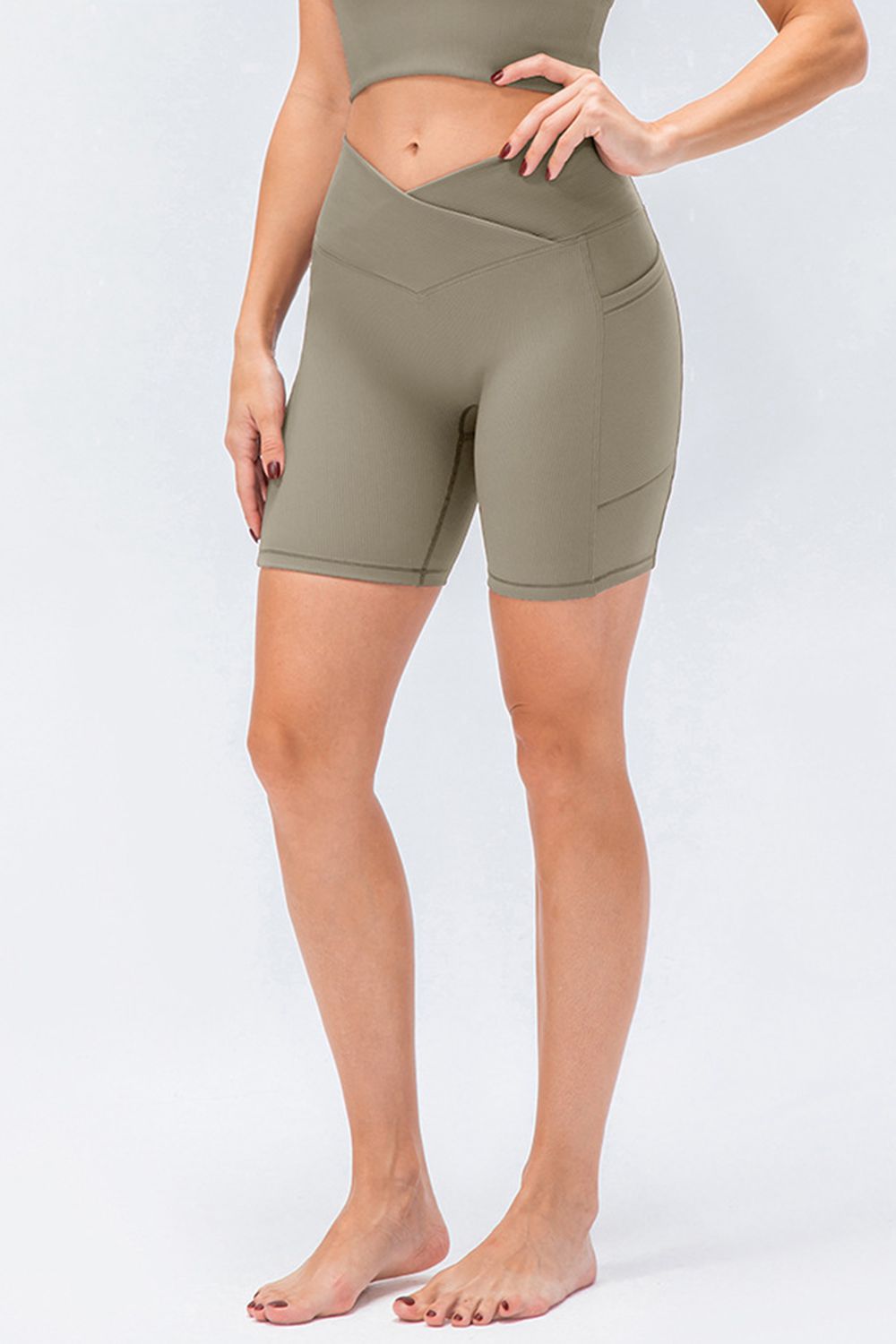 Model wears taupe slim-fit sports shorts with a high V-waistband, stretchy biker-length silhouette, flat seams, and a side pocket while standing with one hand on her hip.