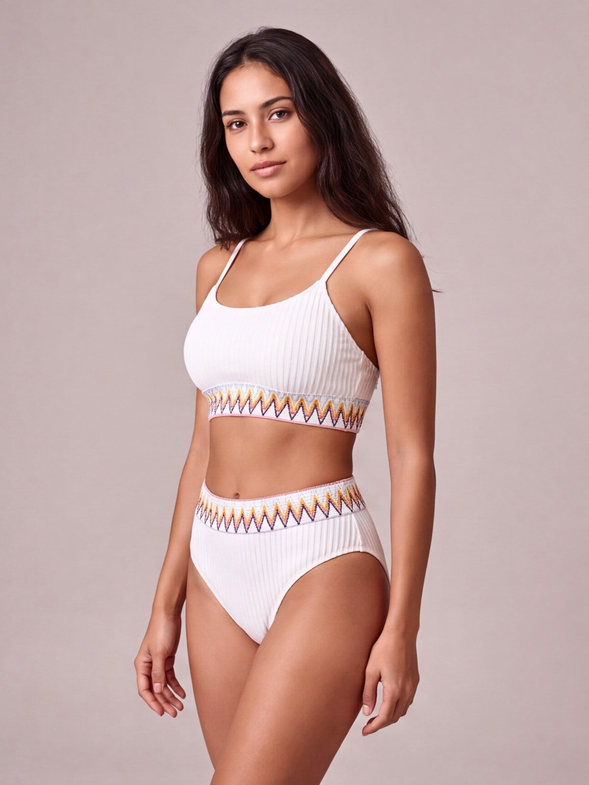 A model poses in a white textured high-waist bikini with thin straps, vertical ribbing, and colorful contrast zigzag banding on the top and bottoms.