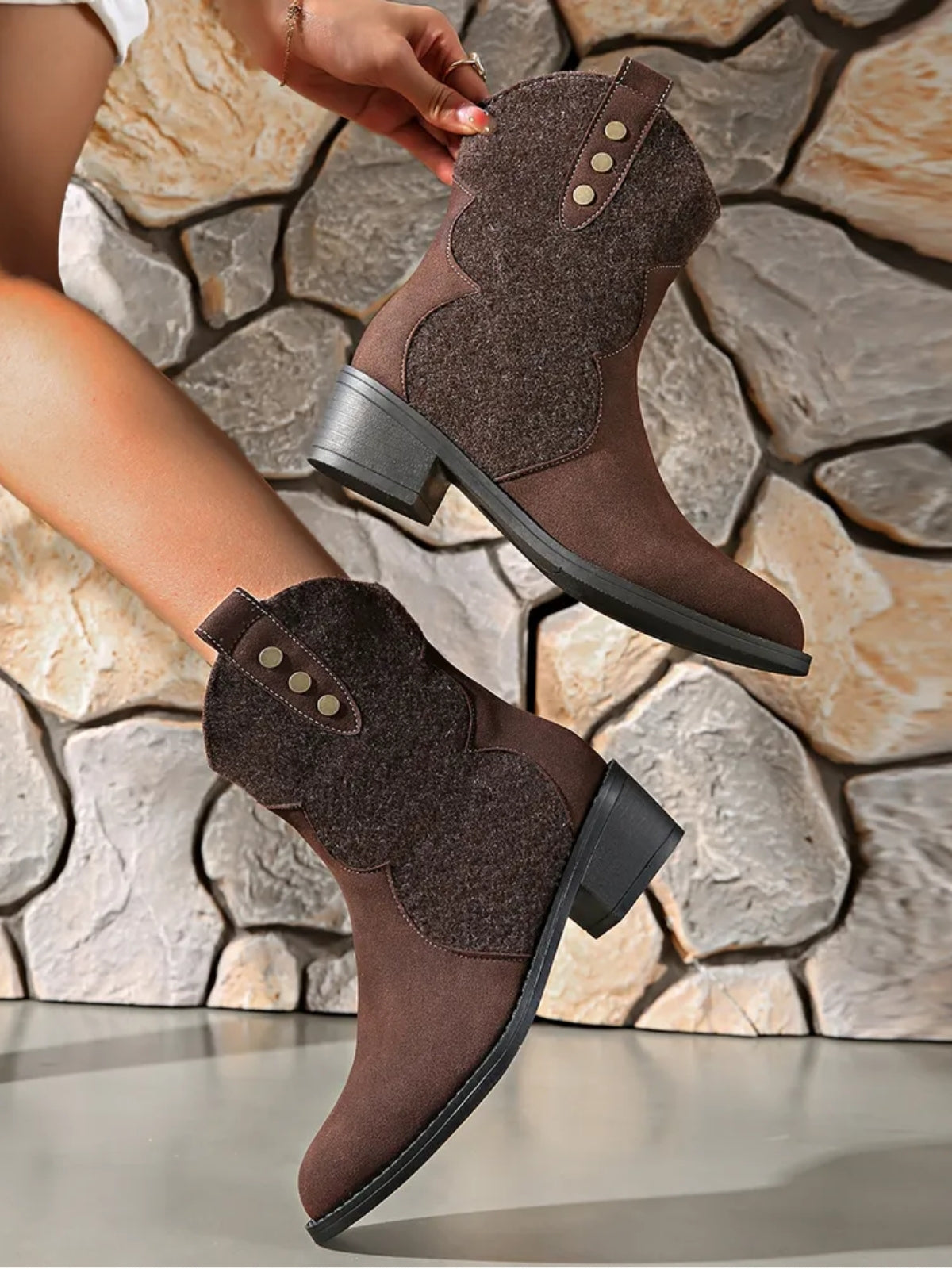 A model displays brown suede point-toe boots with curved panel construction, decorative three-button straps, and low block heels.