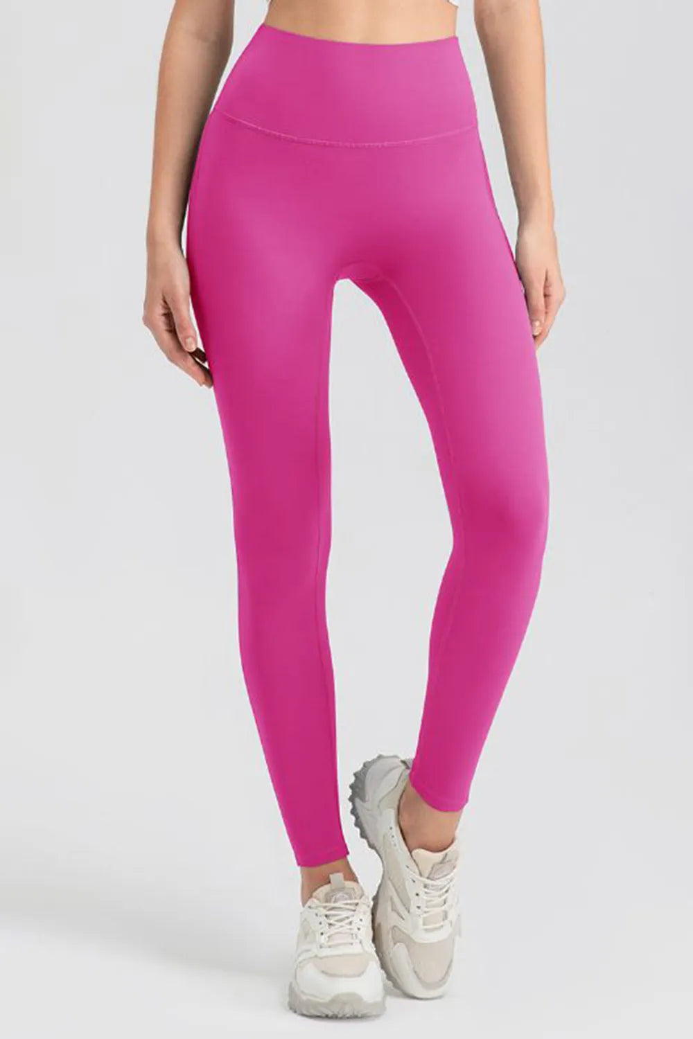 Model wearing bright pink high-waist skinny active pants with a smooth, stretchy legging silhouette, ankle-length fit, and crossed-leg pose.