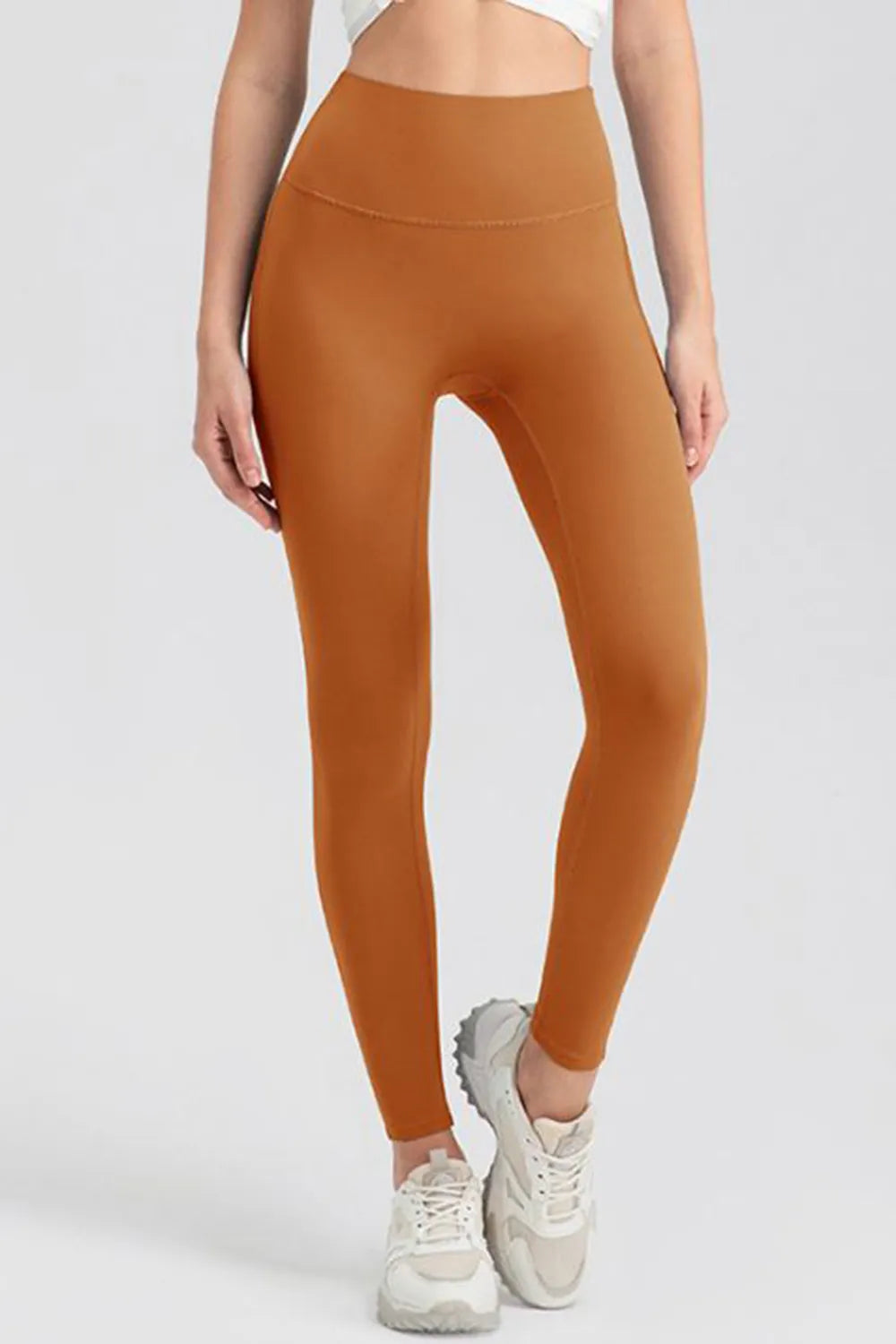 Model wearing high-waist rust orange skinny active leggings with a smooth fitted silhouette, wide waistband, and full-length construction while standing with one leg crossed.