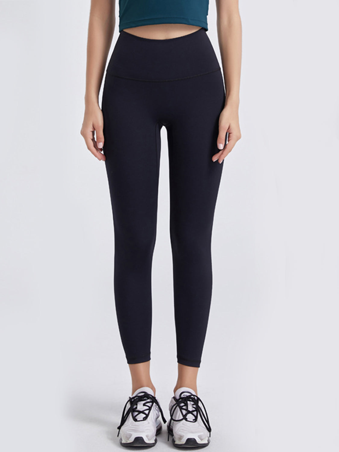 Model stands facing forward in black sports leggings with a wide waistband, high-rise fit, ankle-length tapered silhouette, and visible curved waistband seam.