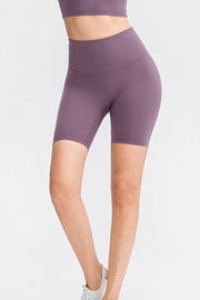 Model wears solid purple slim-fit sports shorts with a wide waistband, smooth construction, and a fitted mid-thigh silhouette while standing with legs crossed.