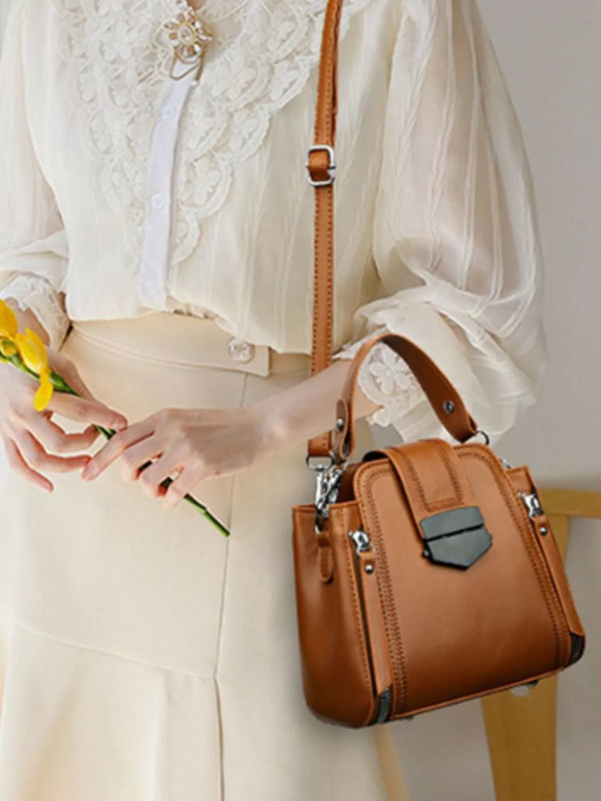 Model in a white lace-trim blouse holds a yellow flower while wearing a medium brown PU leather crossbody bag with a structured silhouette, front pocket, zippers, and buckle details.