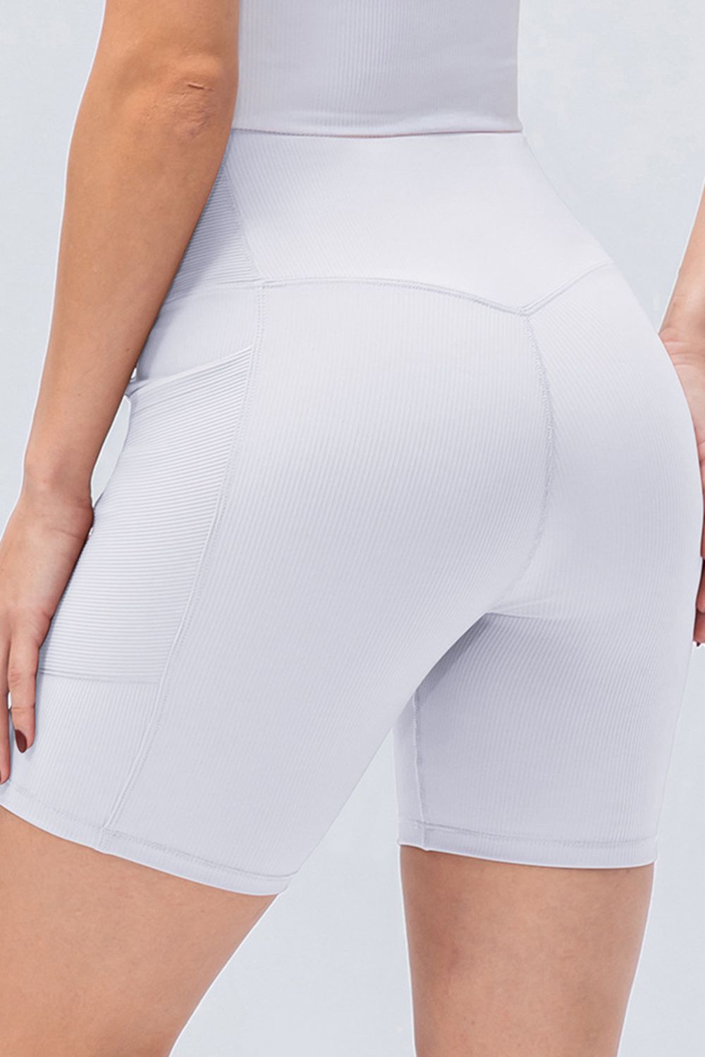 A model wears white slim-fit sports shorts with a V-waistband, ribbed stretch fabric, a side pocket, and visible panel seams in a close-up pose.