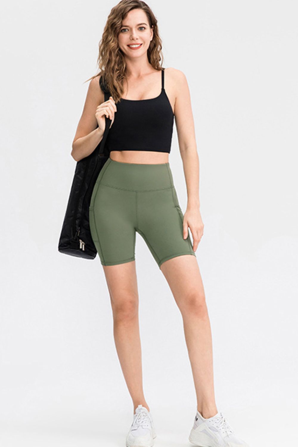 A model wears olive-green slim-fit sports shorts with a wide waistband, fitted biker-length silhouette, and visible side pockets while standing in a relaxed pose.
