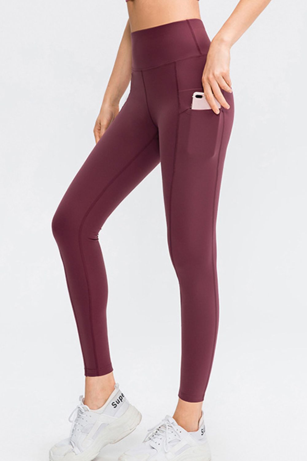 Model wearing solid burgundy slim-fit long sports leggings with a wide waistband, side pocket, and seamed construction, posed with one knee bent.
