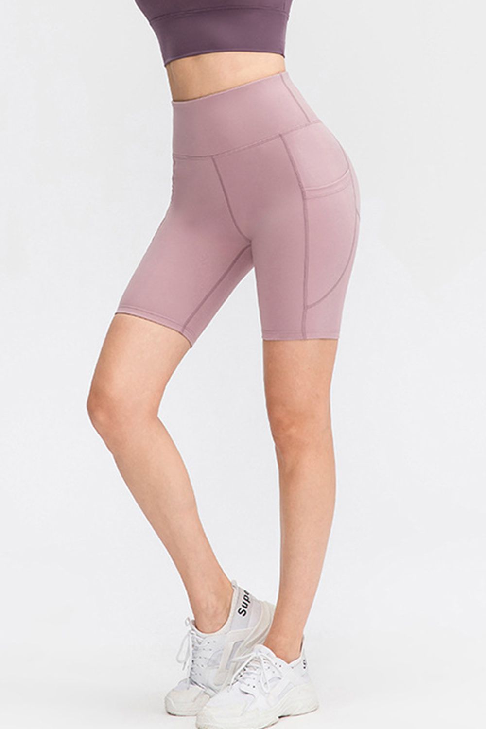 Model wears mauve wide-waistband sports shorts with a fitted knee-length silhouette, curved pocket panels, and visible seam construction while standing with crossed legs.