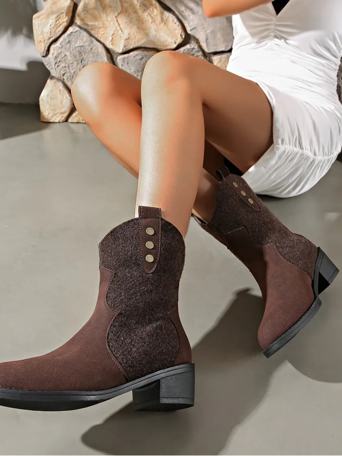 Model seated in a white dress wears brown suede point-toe boots with contrasting panels, decorative button tabs, stitched construction, and low block heels.