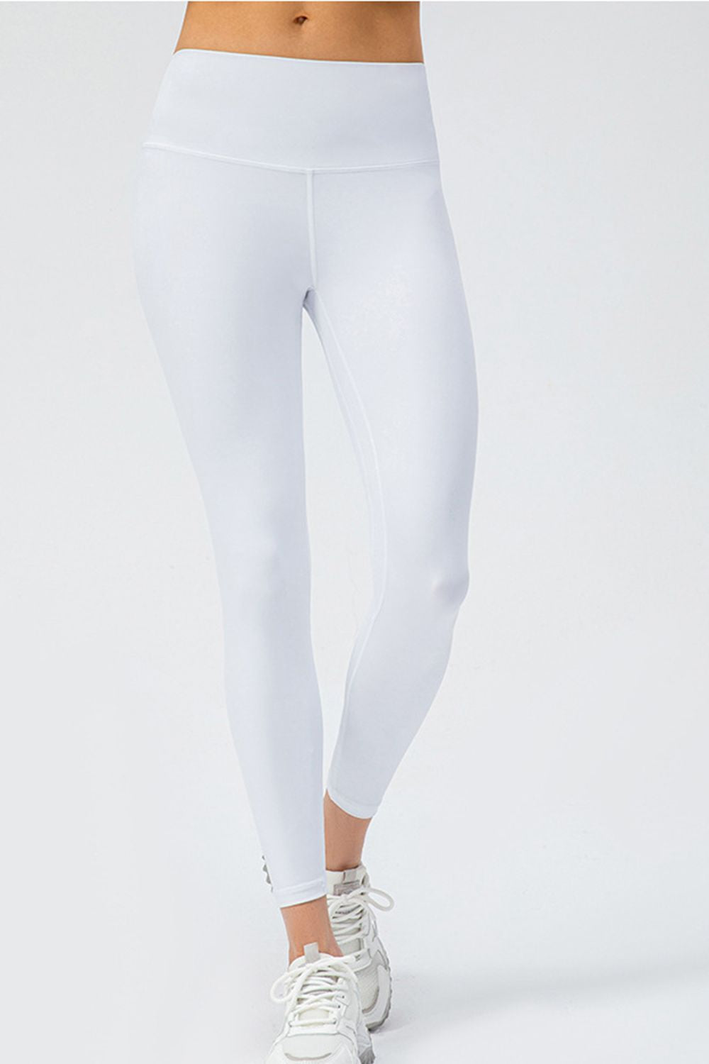 Model wears solid white slim-fit active leggings with a wide waistband, ankle-length silhouette, and visible front seams while standing with one leg crossed.