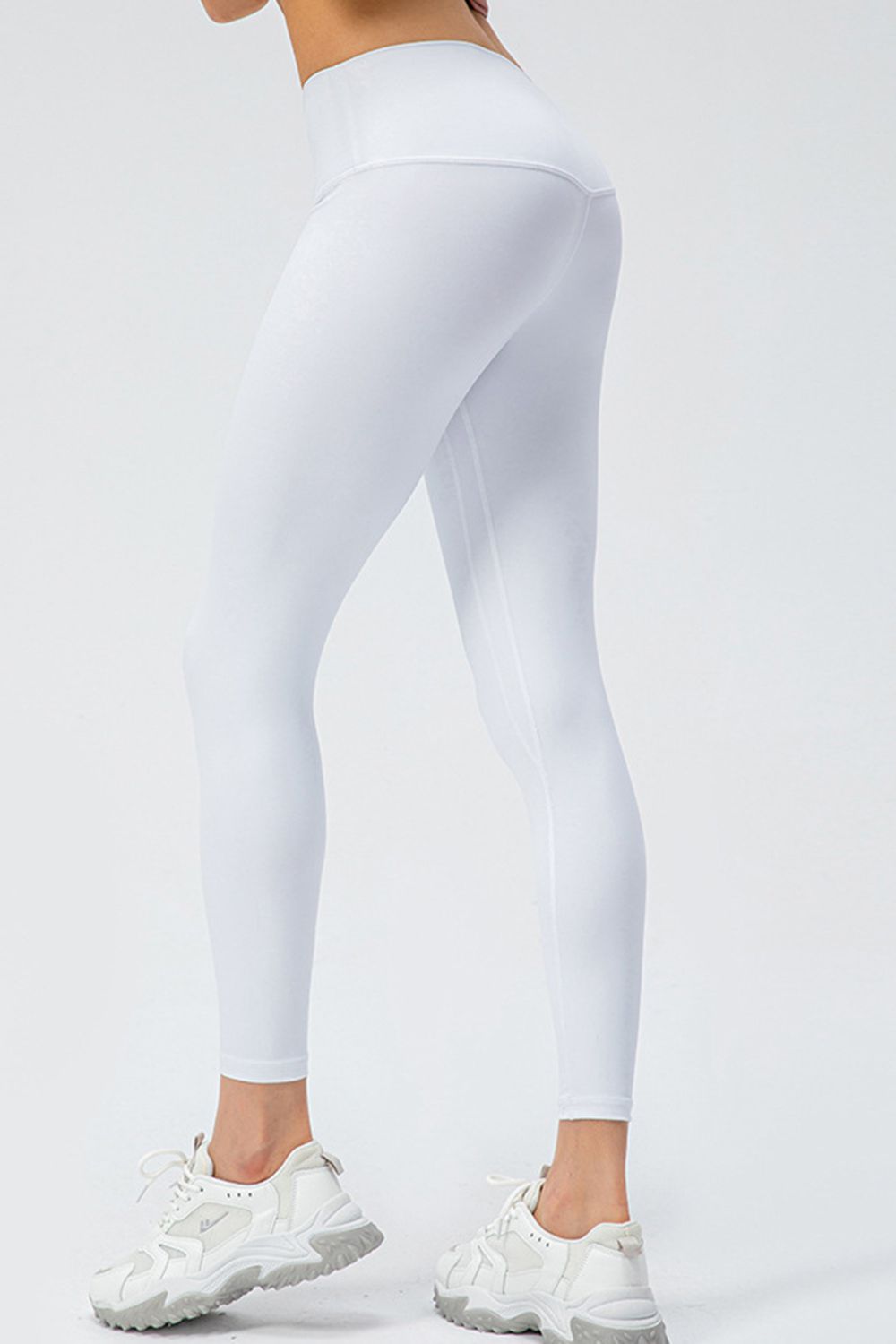 A model poses in white wide-waistband active leggings, showing a slim ankle-length fit, stretchy silhouette, and curved seams through the hips and legs.