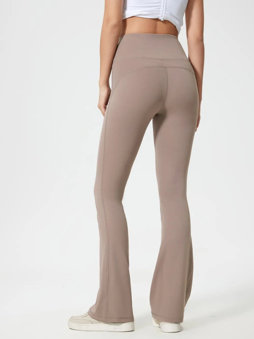 Model shown from behind wearing taupe high-waist active pants with a smooth, close-fitting silhouette, curved seam details, and subtly flared legs.