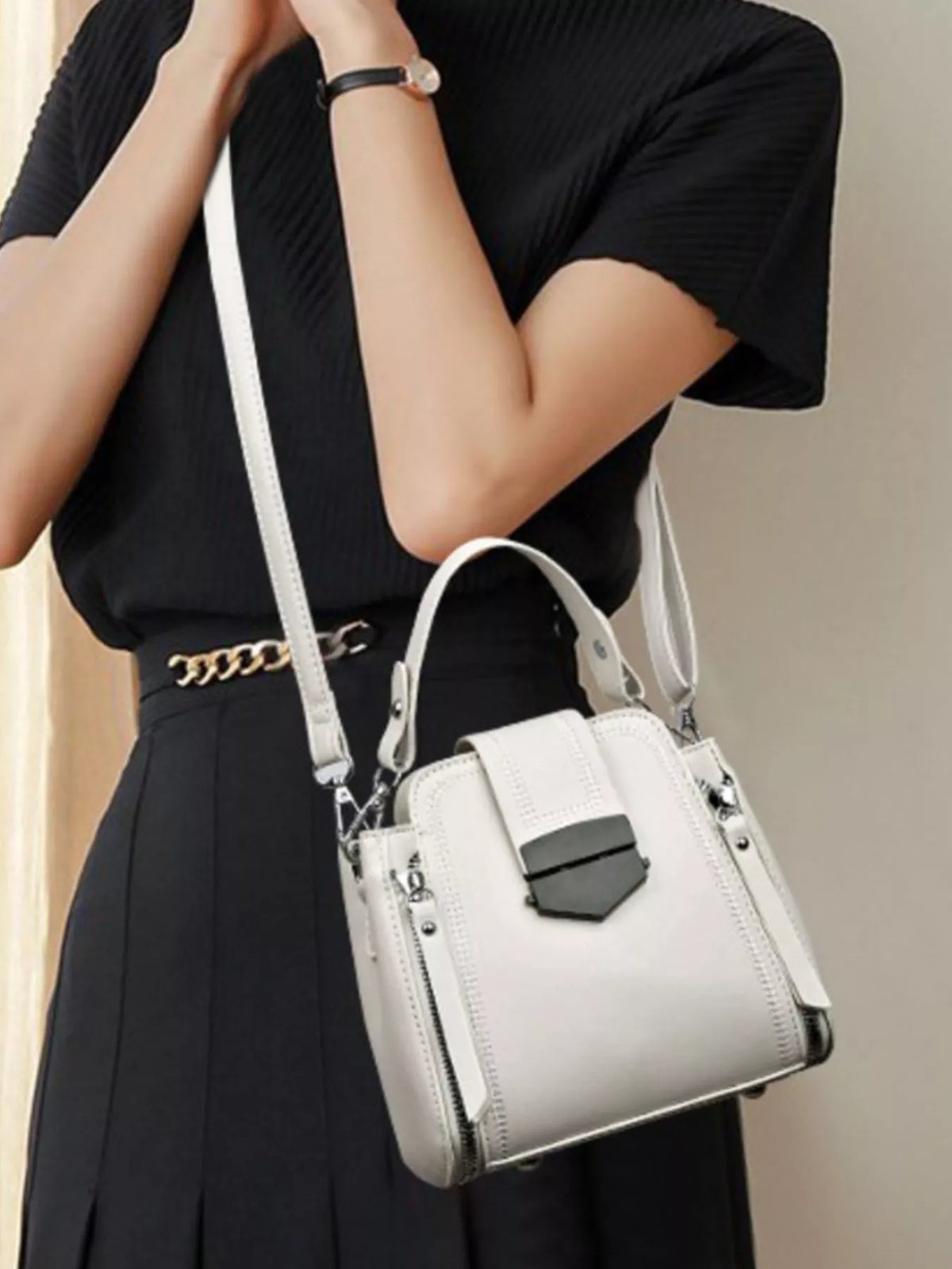 A model holds a medium white PU leather crossbody bag with a structured bucket silhouette, stitched panels, dual straps, side zippers, and a dark front clasp.
