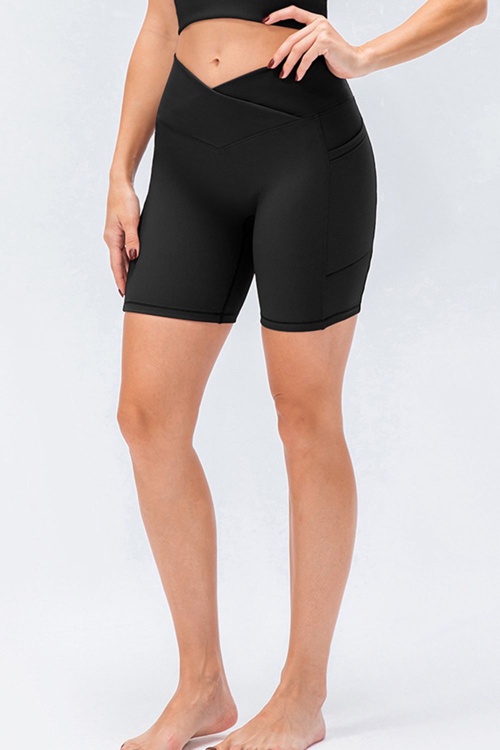 A standing model wears solid black slim-fit sports shorts with a V-shaped waistband, fitted above-knee silhouette, and visible panel seams, posing with one hand on her hip.