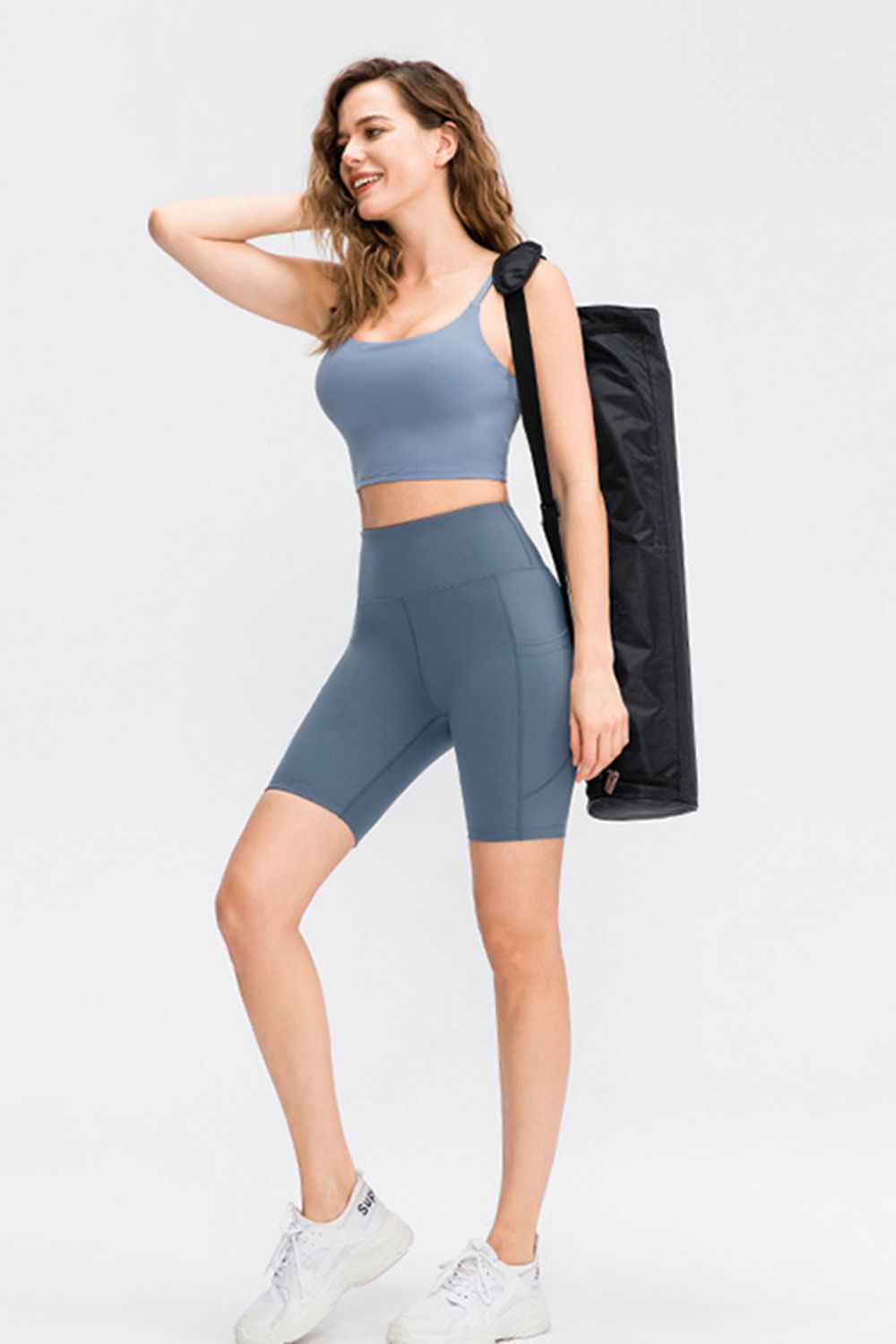 Model wears solid blue-gray sports shorts with a wide waistband, fitted mid-thigh silhouette, stretch construction, and visible side pockets while posing with one leg bent.
