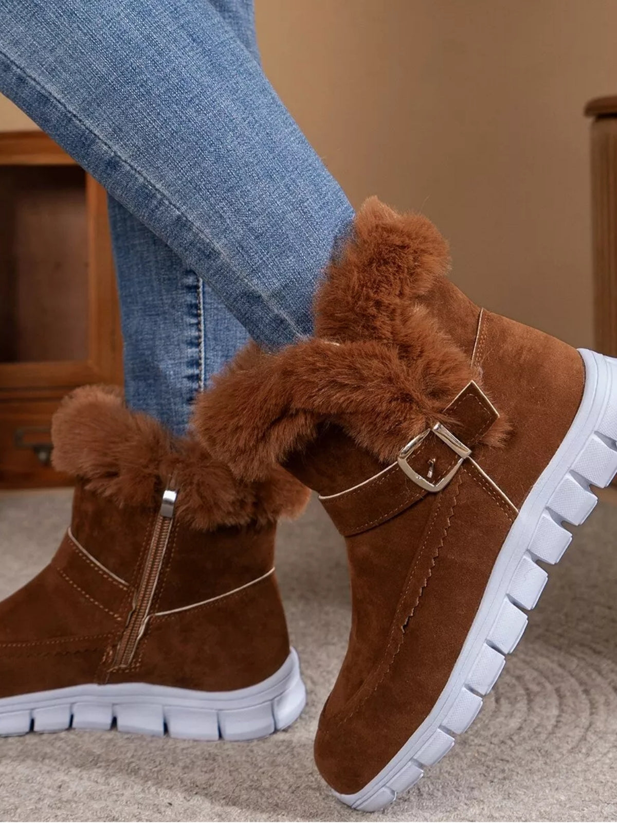 Brown suede fuzzy-trim buckle boots with flat white rubber lug soles are shown on crossed legs, featuring side zippers and warm plush cuffs.