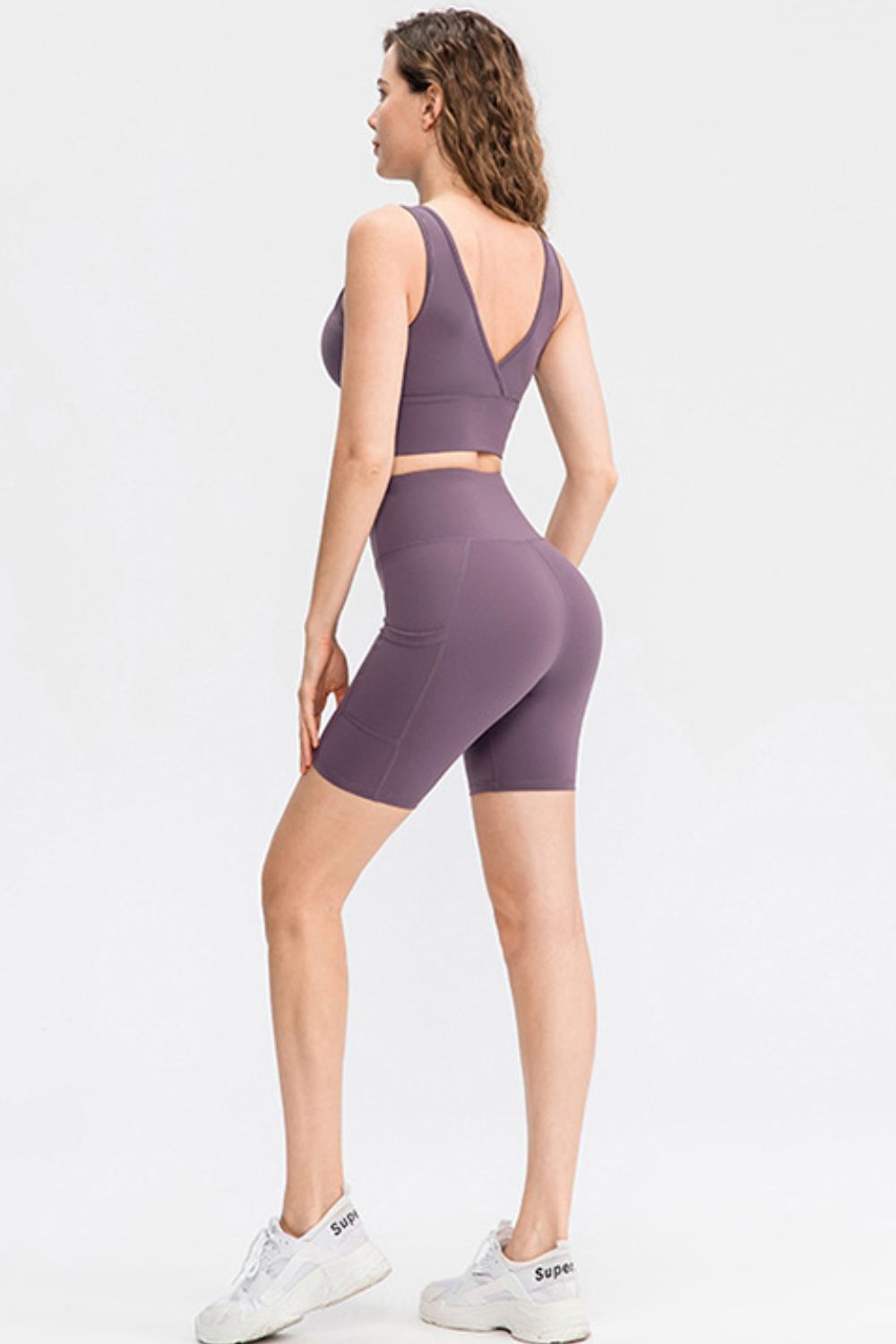 A model poses from behind in solid purple sporty shorts with a wide waistband, slim-fit silhouette, moderate stretch, and a visible side pocket.