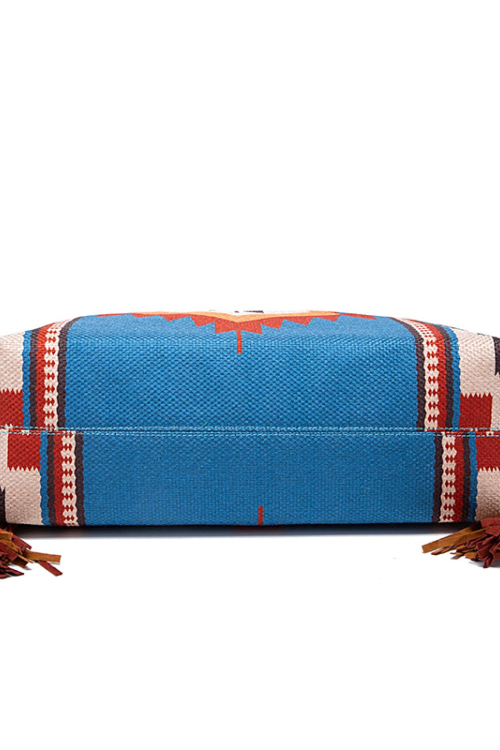 Close-up of an oversize geometric canvas tote bag in blue with red, cream, and black patterned panels, visible stitching, and fringe trim.